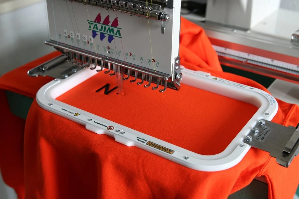 Macchina da ricamo Tajima in azione mentre personalizza una t-shirt arancione con iniziale
